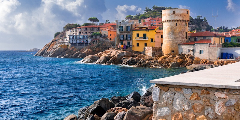 The perfect tiny seaside village of "Giglio Porto" with multi colored houses, an ancient defensive tower and a rocky coastline against a deep blue Mediterranean sea. - Giglio Island, Tuscany, Italy