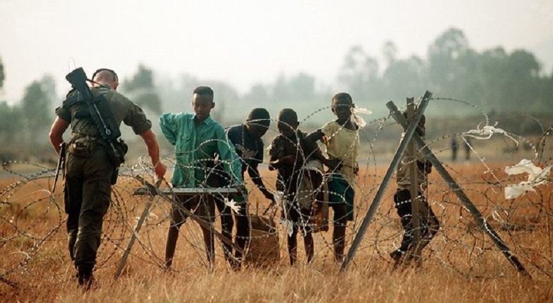 A French soldier, one of the international force supporting the relief effort adjusts the concertina wire surrounding the airport while Rwandan refugee children watch.