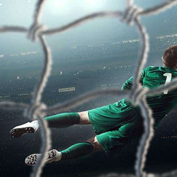 Behind the gate net view - a male soccer goalie jumping in motion for a ball while defending his gates on wide angle panoramic image of a outdoor soccer stadium or arena full of spectators under a sunny sky. The image has depth of field with the focus on the foreground part of the pitch. Players is wearing unbranded soccer uniform.