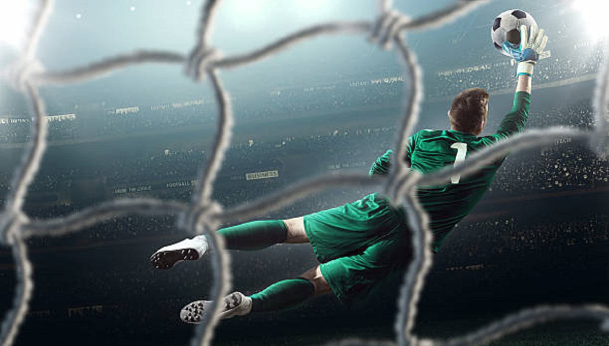 Behind the gate net view - a male soccer goalie jumping in motion for a ball while defending his gates on wide angle panoramic image of a outdoor soccer stadium or arena full of spectators under a sunny sky. The image has depth of field with the focus on the foreground part of the pitch. Players is wearing unbranded soccer uniform.